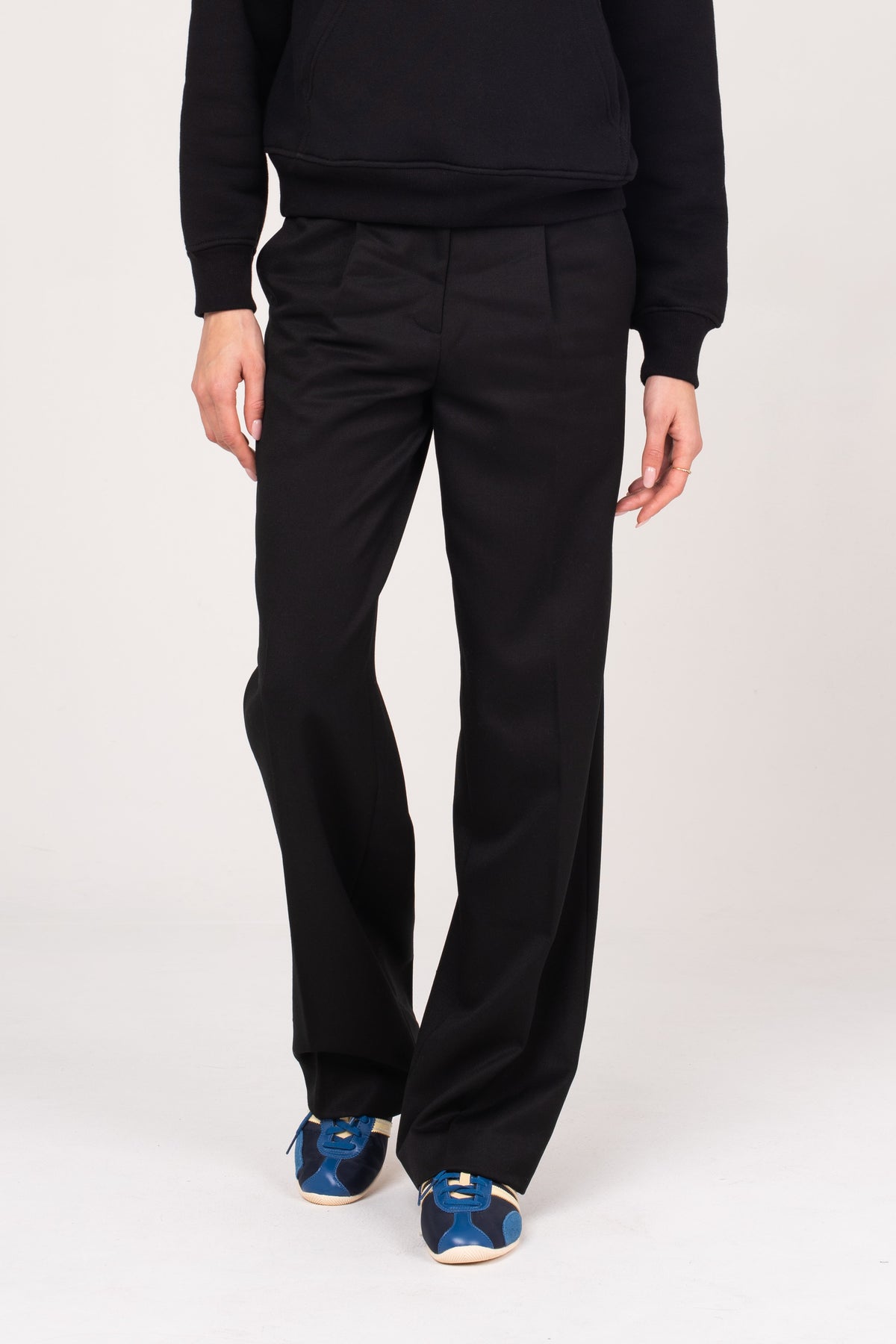 A close-up of the NO.29 women's black pleated trousers, styled with a black NO.29 hoodie and blue sneakers. A refined take on classic tailoring, designed for a relaxed yet polished fit.